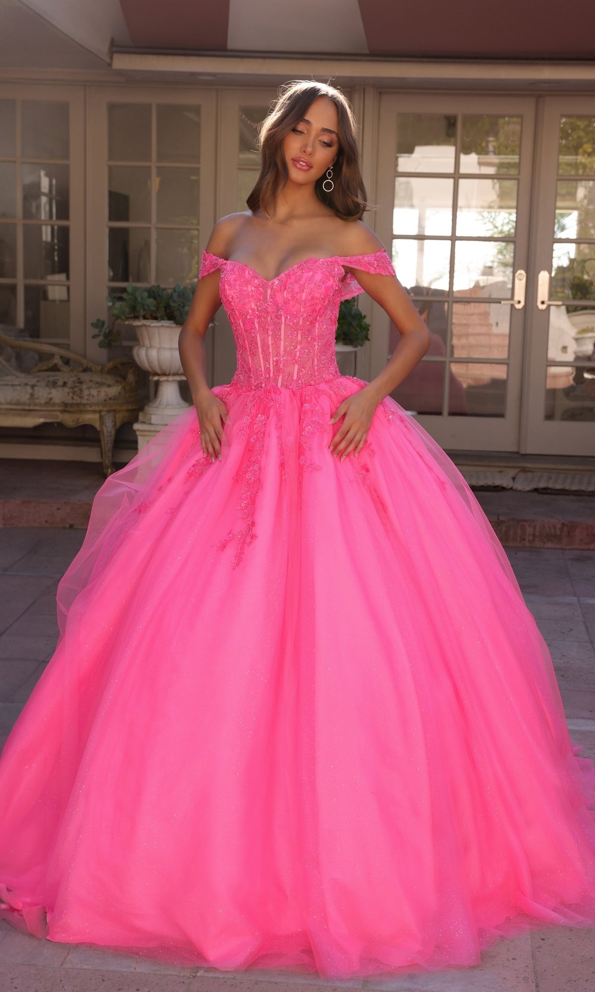 A woman outdoors wears the Bright Off-the-Shoulder Long Prom Ball Gown H1349, featuring a vibrant pink hue, fitted bodice, and full tulle skirt. French doors and outdoor furniture appear in the background.
